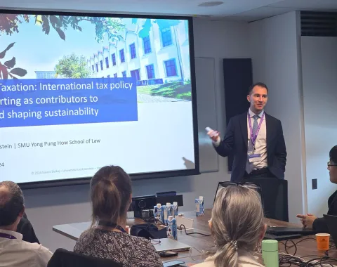 Ein Vortragender steht vor einer Präsentation mit dem Titel "Sustainable Taxation: International tax policy and reporting as contributors to achieving and shaping sustainability". Im Raum sitzen mehrere Personen, die dem Vortrag folgen.
