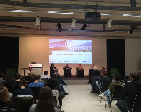 Ein Auditorium mit mehreren Personen, die einer Podiumsdiskussion zuhören. Drei Redner sitzen auf der Bühne vor einer Leinwand mit der Aufschrift „Discussion: 'Micro-credentials are nothing new'“. Die Veranstaltung findet in einem modernen Konferenzraum statt.