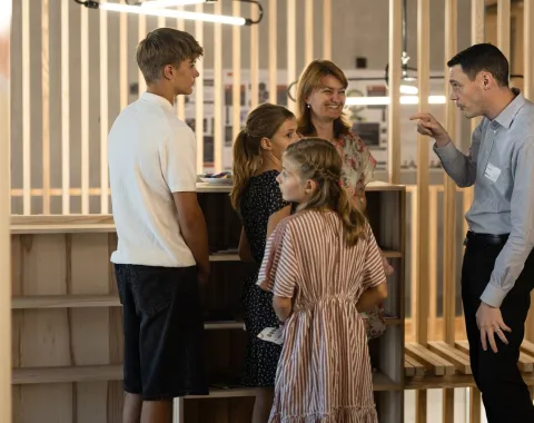 Eine Gruppe von Menschen steht in einem modernen Raum mit Holzelementen. Ein Mann spricht gestikulierend mit einer Frau und drei Kindern. Die Szene vermittelt eine freundliche und einladende Atmosphäre während des Tags der offenen Tür an der Universität Liechtenstein.