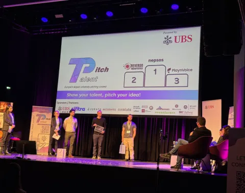 Eine Bühne mit vier Personen, die an einem Pitch-Wettbewerb teilnehmen. Im Hintergrund ist eine Leinwand mit dem Logo von "Talent Pitch" und den Namen der Start-ups "Reverso Therapeutics", "nepsos" und "PlaynVoice" zu sehen. Das Event wird von UBS gesponsert. Zuschauer sitzen auf der rechten Seite.