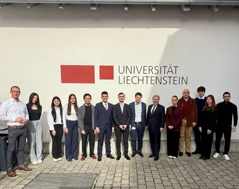 Eine Gruppe von Menschen steht vor einer Wand mit dem Logo der Universität Liechtenstein. Sie sind formell gekleidet und lächeln in die Kamera.