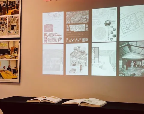 Ein Ausstellungstisch mit offenen Büchern steht vor einer Wand, auf der Architekturzeichnungen und Pläne projiziert sind. Links sind Fotos von Designprojekten zu sehen. Oben leuchtet ein "swisstech" Schild in roter Schrift.