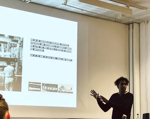 Ein Vortragender steht vor einer Projektionsleinwand und präsentiert Inhalte zu urbanem Design. Auf der Leinwand sind Text und Bilder zu sehen. Im Vordergrund sitzen Zuhörer. Der Raum wirkt modern und technisch ausgestattet.