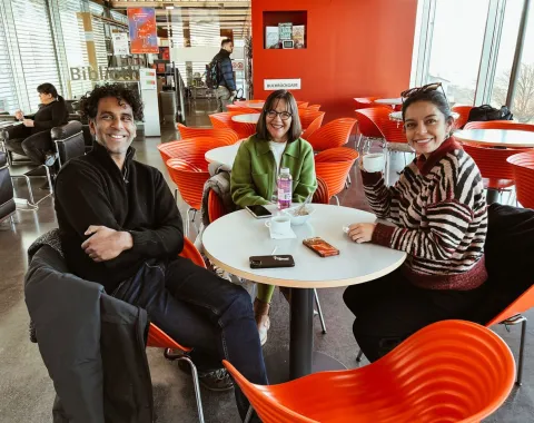Drei Personen sitzen lächelnd an einem Tisch in einem modernen Café mit roten Stühlen. Sie halten Getränke in der Hand und wirken entspannt. Im Hintergrund sind weitere Gäste und eine Bücherwand zu sehen.