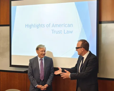 Zwei Männer in Anzügen stehen vor einer Präsentation mit dem Titel "Highlights of American Trust Law". Einer der Männer spricht gestikulierend. Im Hintergrund ist ein Projektionsbildschirm zu sehen.