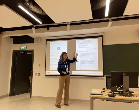 Eine Person präsentiert in einem modernen Seminarraum vor einer Leinwand mit zwei projizierten Dokumenten. Im Vordergrund steht ein Tisch mit einem Computerbildschirm.