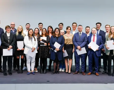 Eine Gruppe von Absolventinnen und Absolventen steht auf einer Bühne und hält Zertifikate in den Händen. Sie lächeln in die Kamera. Die Personen sind formell gekleidet, und im Hintergrund ist eine neutrale Wand zu sehen.
