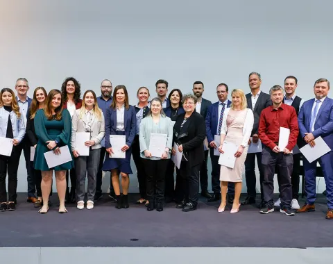 Eine Gruppe von Absolventinnen und Absolventen steht auf einer Bühne und hält Zertifikate in den Händen. Sie lächeln in die Kamera. Die Personen sind formell gekleidet, und im Hintergrund ist eine neutrale Wand zu sehen.