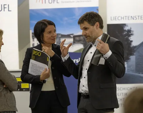 Drei Personen stehen in einem Gespräch vor Roll-Up-Bannern der Universität Liechtenstein und des Liechtenstein-Instituts. Eine Person hält ein Buch in der Hand. Die Szene findet in einem formellen Rahmen statt, vermutlich bei einer akademischen Veranstaltung.