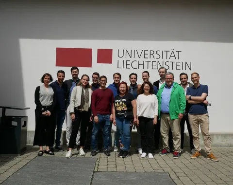 Gruppenfoto von Alumni des Abschlussjahrgangs 2013 des Masters in Banking and Financial Management vor dem Logo der Universität Liechtenstein. Die Personen stehen lächelnd zusammen auf dem Campus bei sonnigem Wetter.