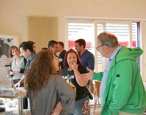 Eine Gruppe von Menschen unterhält sich lebhaft in einem hellen Raum auf dem Campus der Universität Liechtenstein. Im Hintergrund sind Fenster und ein Plakat zu sehen.