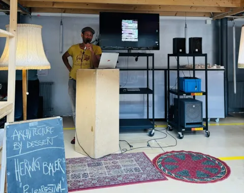 Ein Mann steht an einem Rednerpult in einem Raum mit Holzdach. Neben ihm ist ein Bildschirm mit einer Präsentation und ein Schild mit der Aufschrift „Architecture by Dessert, Herwig Bauer, Poolbarfestival“. Der Raum ist mit Teppichen und Stehlampen dekoriert.