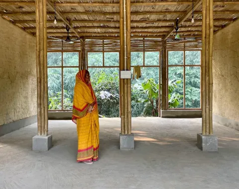 Eine Frau in traditioneller indischer Kleidung steht in einem offenen Raum mit Lehmwänden und Bambussäulen. Große Fenster im Hintergrund bieten einen Blick auf üppiges Grün.
