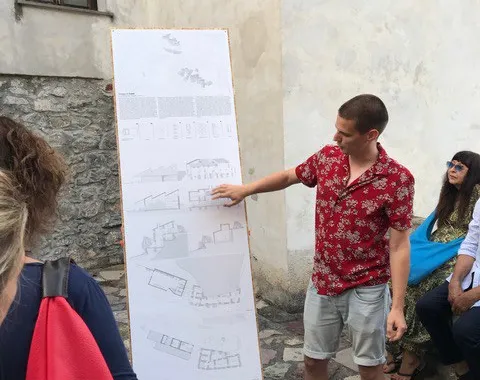 Ein Student präsentiert architektonische Skizzen auf einem großen Plakat in einem historischen Dorf. Um ihn herum stehen und sitzen mehrere Personen, die aufmerksam zuhören. Die Szene spielt sich in einem gepflasterten Außenbereich ab.