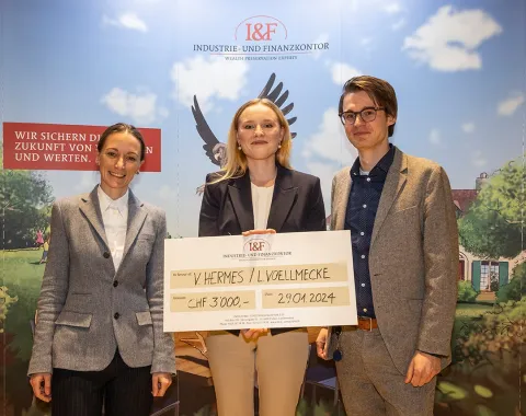 Drei Personen stehen lächelnd vor einem Hintergrund mit dem Logo der Industrie- und Finanzkontor. Eine Person hält einen großen Scheck mit der Aufschrift "CHF 3,000" und einem Datum. Im Hintergrund ist ein stilisiertes Bild mit einem Adler und einem Haus zu sehen.