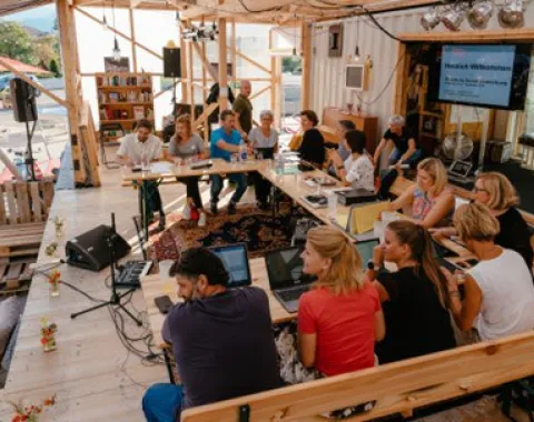 Eine Gruppe von Menschen sitzt in einem offenen, modularen Raum an einem Tisch mit Laptops. Der Raum ist aus Holz und enthält Bücherregale und technische Ausrüstung. Es herrscht eine lebhafte Diskussionsatmosphäre.