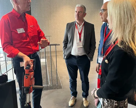 Eine Gruppe von vier Personen steht in einem modernen Gebäude. Eine Person hält ein Werkzeug und erklärt etwas den anderen. Alle tragen Namensschilder und hören aufmerksam zu.