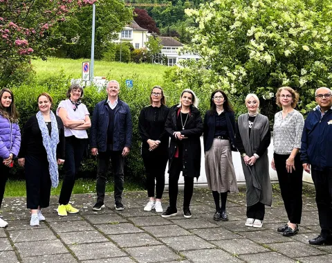 Eine Gruppe von zehn Personen steht draußen auf einem gepflasterten Weg vor einer grünen Hecke und blühenden Bäumen. Die Personen sind in Freizeitkleidung und lächeln in die Kamera. Im Hintergrund sind ein Gebäude und eine Wiese zu sehen.