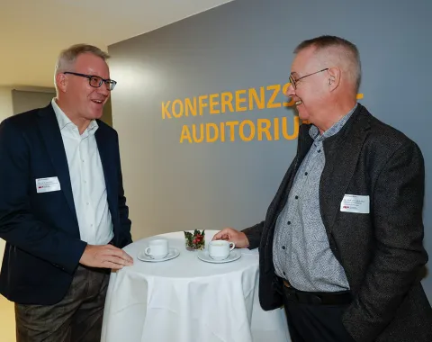 Zwei Männer im Gespräch bei einer Veranstaltung an der Universität Liechtenstein. Sie stehen an einem Stehtisch mit Kaffeetassen im Bereich "Konferenzzentrum Auditorium".
