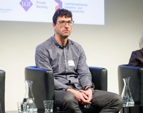 Eine Person sitzt auf einer Bühne in einem schwarzen Sessel. Im Hintergrund sind Logos von Sponsoren oder Partnern zu sehen. Auf dem Tisch vor der Person stehen Wasserkaraffen und Gläser.