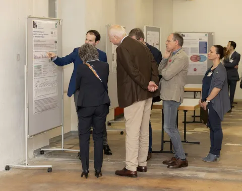 Eine Gruppe von Personen betrachtet eine wissenschaftliche Posterausstellung in einem Innenraum. Ein Mann zeigt auf ein Poster und erklärt den Inhalt. Im Hintergrund sind weitere Poster und Personen zu sehen.
