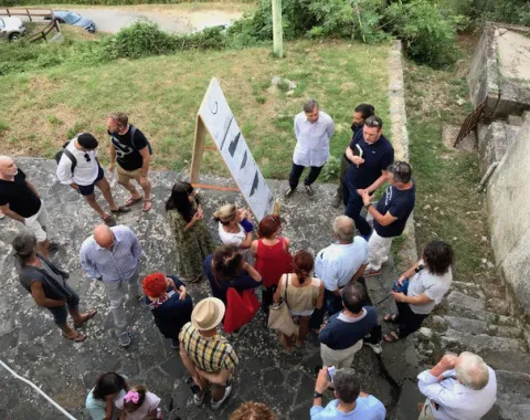 Eine Gruppe von Menschen steht im Freien um eine Tafel mit Bildern oder Plänen. Die Szene findet auf einem gepflasterten Platz mit angrenzendem Grünbereich statt. Die Personen scheinen in eine Diskussion oder Präsentation vertieft zu sein.