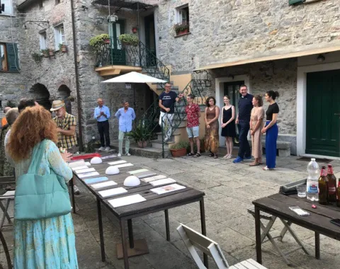 Eine Gruppe von Menschen versammelt sich in einem historischen Innenhof eines Dorfes. Auf Tischen liegen Dokumente und Objekte aus. Die Szene zeigt eine Präsentation oder Diskussion im Freien, umgeben von alten Steinmauern und Pflanzen.