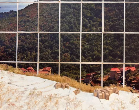 Ein Modell zeigt eine hügelige Landschaft mit kleinen Gebäuden und Vegetation, im Hintergrund ist ein großes Foto einer bewaldeten Berglandschaft zu sehen.