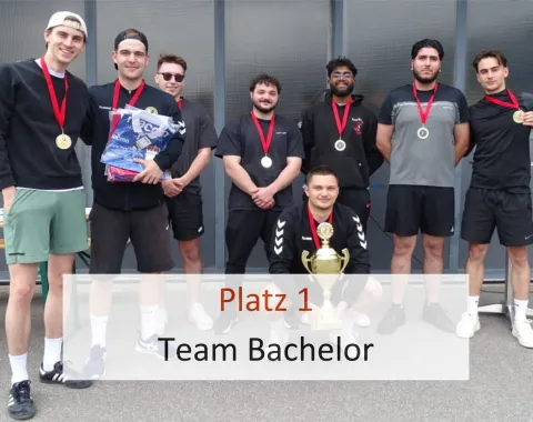 Acht junge Männer stehen in sportlicher Kleidung vor einer Wand. Sie tragen Medaillen um den Hals. Einer kniet vorne und hält einen Pokal. Text im Bild: "Platz 1 Team Bachelor".