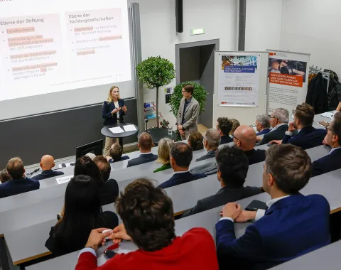 Ein Vortrag in einem Hörsaal der Universität Liechtenstein: Eine Person präsentiert vor einer Gruppe von Zuhörern, die in gestaffelten Sitzreihen sitzen. Im Hintergrund sind eine Leinwand mit Präsentationsfolien und ein Roll-up-Banner zu sehen.