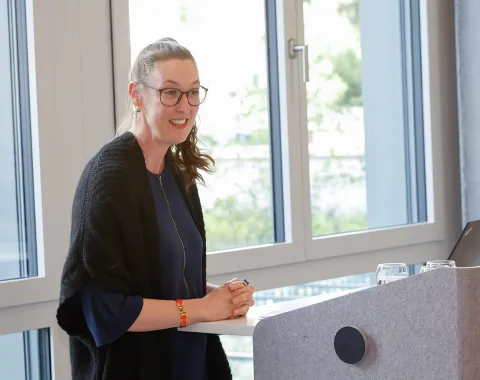 Eine Person steht lächelnd an einem Rednerpult in einem modernen Raum mit großen Fenstern. Sie trägt eine Brille und hält einen Stift in der Hand. Im Hintergrund sind Bäume zu sehen.