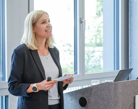 Eine Person hält einen Vortrag an einem Rednerpult in einem modernen Raum mit großen Fenstern. Sie trägt einen dunklen Blazer und hält ein Mikrofon sowie Notizen in der Hand.