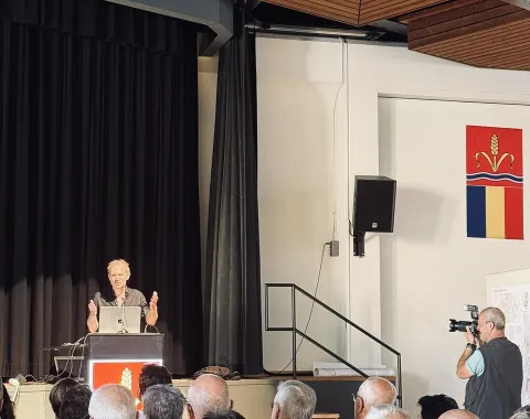 Ein Vortragender steht an einem Rednerpult vor einem Publikum in einem Saal. Im Hintergrund sind schwarze Vorhänge und ein Fotograf macht Bilder. An der Wand hängt ein Wappen.