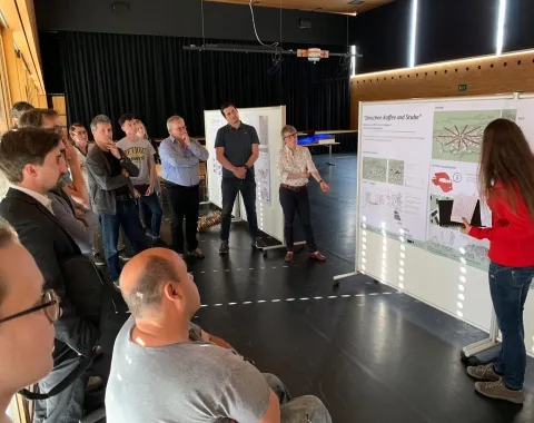 Mehrere Menschen bei einer Präsentation einer Studentin mit Architekturprojekten an einer Pinnwand