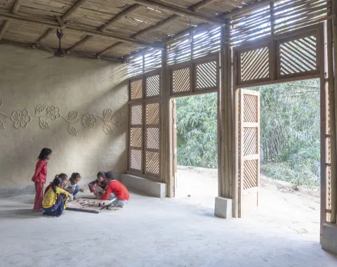Kinder spielen auf dem Boden eines offenen, aus Naturmaterialien gebauten Raums mit Blick ins Grüne.