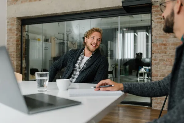Zwei Männer in einem modernen Büro unterhalten sich an einem Tisch. Der Mann im Vordergrund schreibt in ein Notizbuch, während der andere Mann, der zurückgelehnt und lächelnd ist, zuhört. Auf dem Tisch stehen ein Laptop, ein Glas Wasser und eine Tasse. Im Hintergrund sind Glaswände und Ziegelwände zu sehen, die dem Raum eine offene und zeitgemäße Atmosphäre verleihen.