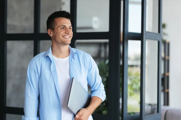 Ein junger Mann lächelt und blickt zur Seite, während er einen Laptop unter dem Arm hält. Er trägt ein hellblaues Hemd über einem weißen T-Shirt und steht in einem modernen Innenraum mit Glaswänden und Pflanzen im Hintergrund. Die Atmosphäre wirkt entspannt und professionell.