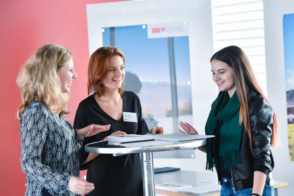 Drei Frauen im Gespräch an einem Stehtisch beim Infotag der Universität Liechtenstein, mit Informationsmaterialien und freundlicher Atmosphäre.