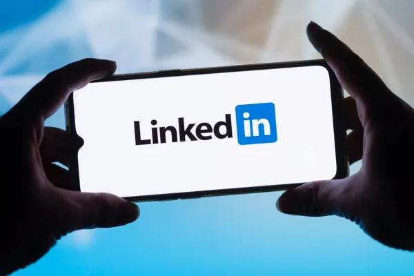 Zwei Hände halten ein Smartphone mit LinkedIn Logo vor einem hellen digitalen Hintergrund mit blauen Lichtreflexen