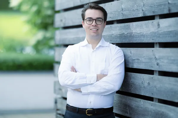 Ein männlicher Studierender im Masterstudiengang Innovative Finance mit Brille und weißem Hemd lehnt lächelnd an einer Holzwand.