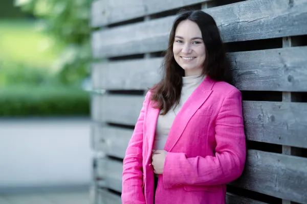 Eine weibliche Studierende im Masterstudiengang Innovative Finance trägt einen auffälligen pinken Blazer und steht entspannt vor einer Holzwand, während sie freundlich in die Kamera lächelt.