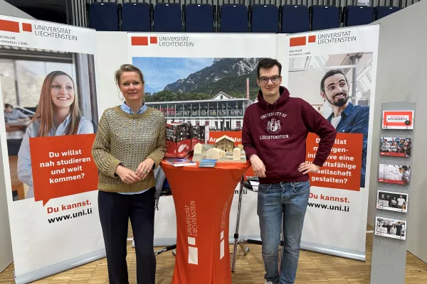 Messestand der Universität Liechtenstein in Düsseldorf mit Infomaterial und zwei Personen vor Informationsbannern.