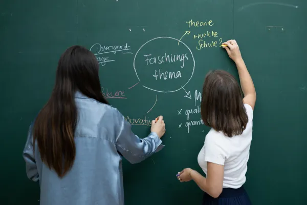 Zwei Studierende entwickeln ein Forschungsthema auf einer beschrifteten grünen Tafel mit Kreide und Farben.