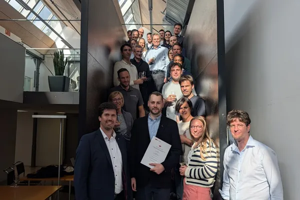 Eine Gruppe von Menschen steht auf einer Treppe in einem modernen Gebäude. Im Vordergrund hält eine Person ein Dokument in der Hand, umgeben von lächelnden Kollegen. Die Szene ist gut beleuchtet durch ein Oberlicht.