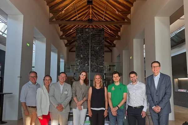 Eine Gruppe von neun Personen steht in einer modernen Halle mit Holzdach und einem Kronleuchter. Sie lächeln in die Kamera. Im Hintergrund sind hohe Fenster und eine Steinwand zu sehen.