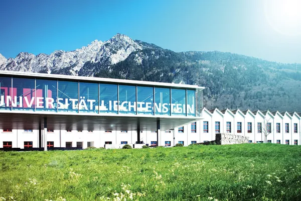 Außenansicht der Universität Liechtenstein mit modernem Glasgebäude und Bergkulisse im Hintergrund, umgeben von grüner Wiese.