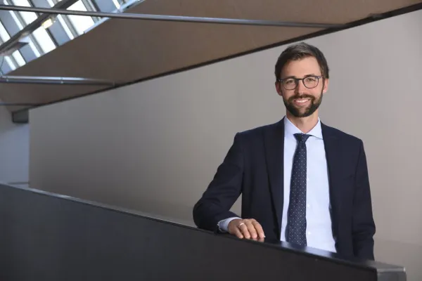 Ein Mann in Anzug und Krawatte steht lächelnd in einem modernen Gebäude mit großen Fenstern.