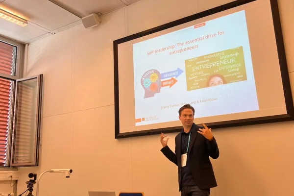 Ein Vortragender präsentiert in einem Seminarraum ein Paper über Selbstführung für Unternehmer. Im Hintergrund ist eine Leinwand mit einer Präsentationsfolie zu sehen. Tische mit Laptops und Kamera sind im Vordergrund.