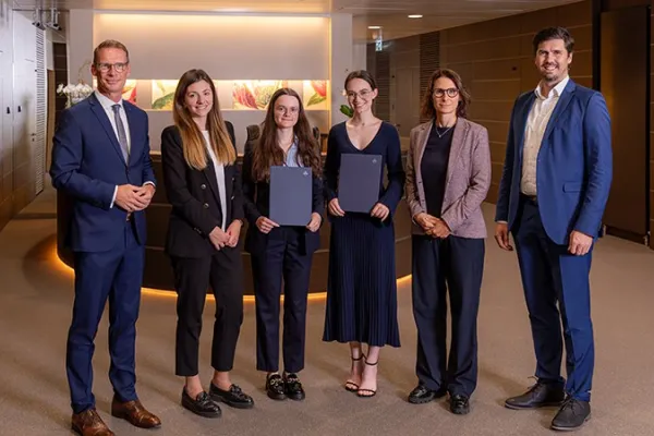 Eine Gruppe von sechs Personen steht in einem modernen Bürogebäude. Zwei Frauen in der Mitte halten Zertifikate und lächeln. Sie werden von vier weiteren Personen flankiert, die ebenfalls lächeln. Die Szene vermittelt eine feierliche Atmosphäre, möglicherweise im Rahmen einer Stipendienverleihung an der Universität Liechtenstein.