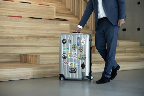 Mann im Anzug zieht silbernen Koffer mit Reiseaufklebern über Campusboden.